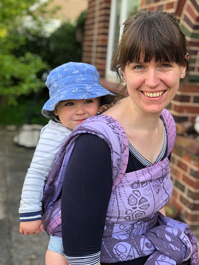 Nelly carries a smiling baby on her back in a purple wrap